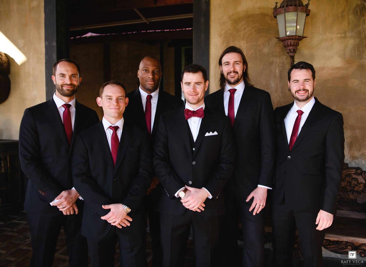 Six groomsmen in black suits with burgundy ties pose together outdoors at a wedding venue.