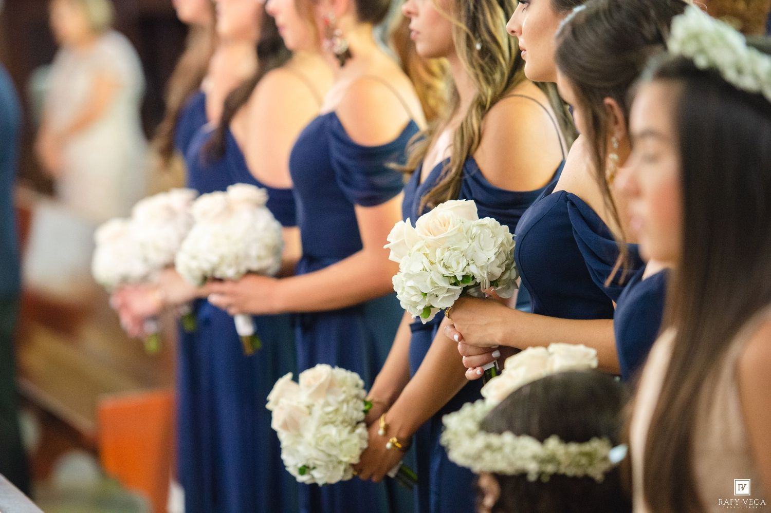 Bridesmaids in navy blue off-shoulder gowns holding white hydrangea bouquets stand in a row during a wedding ceremony.