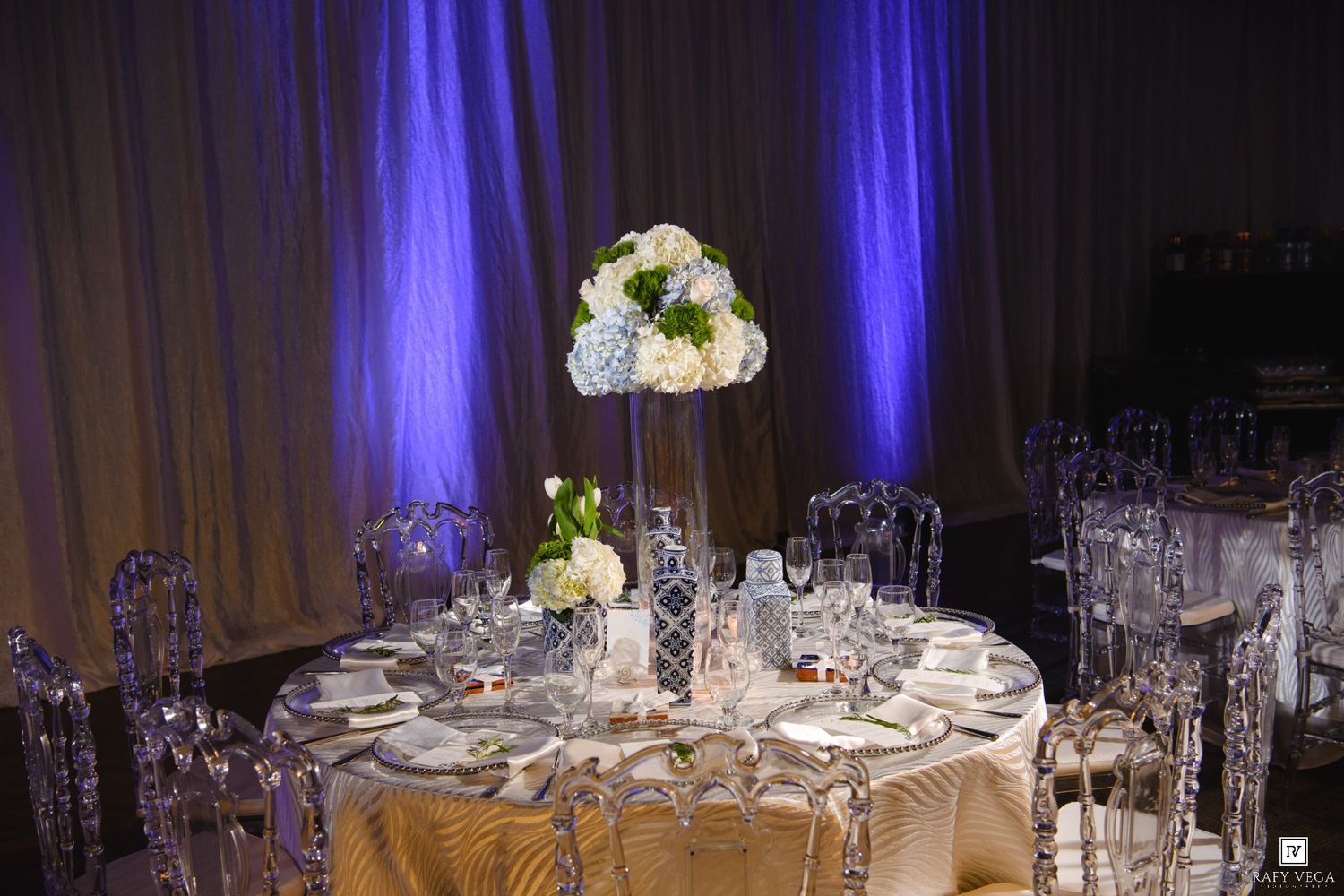 Elegant wedding reception table with tall white floral centerpiece, crystal chairs, and gold linens under blue ambient lighting.
