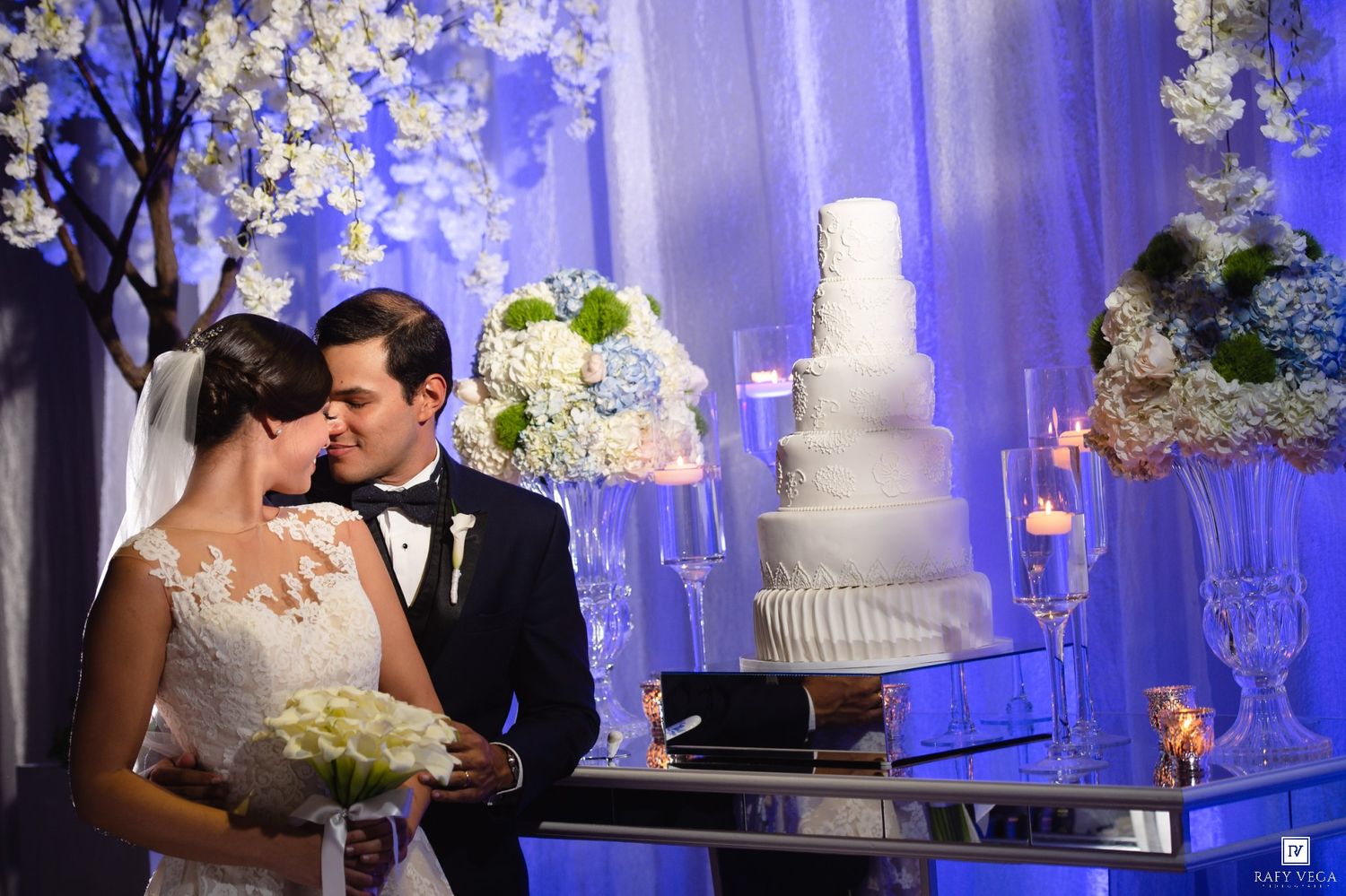 Bride and groom share a romantic moment beside a white tiered wedding cake at an elegant floral-decorated reception.