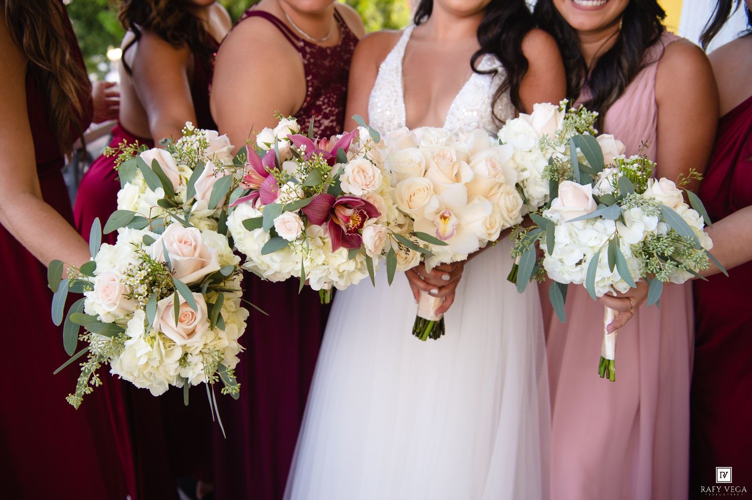 Bride in white gown and bridesmaids in burgundy and blush dresses holding elegant white and pink floral bouquets.