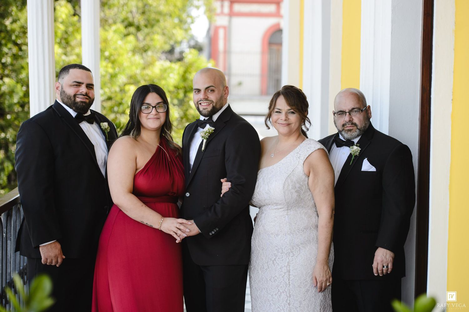 Wedding party posing on a balcony, bride in white gown, bridesmaid in red dress, groom and groomsmen in black tuxedos.