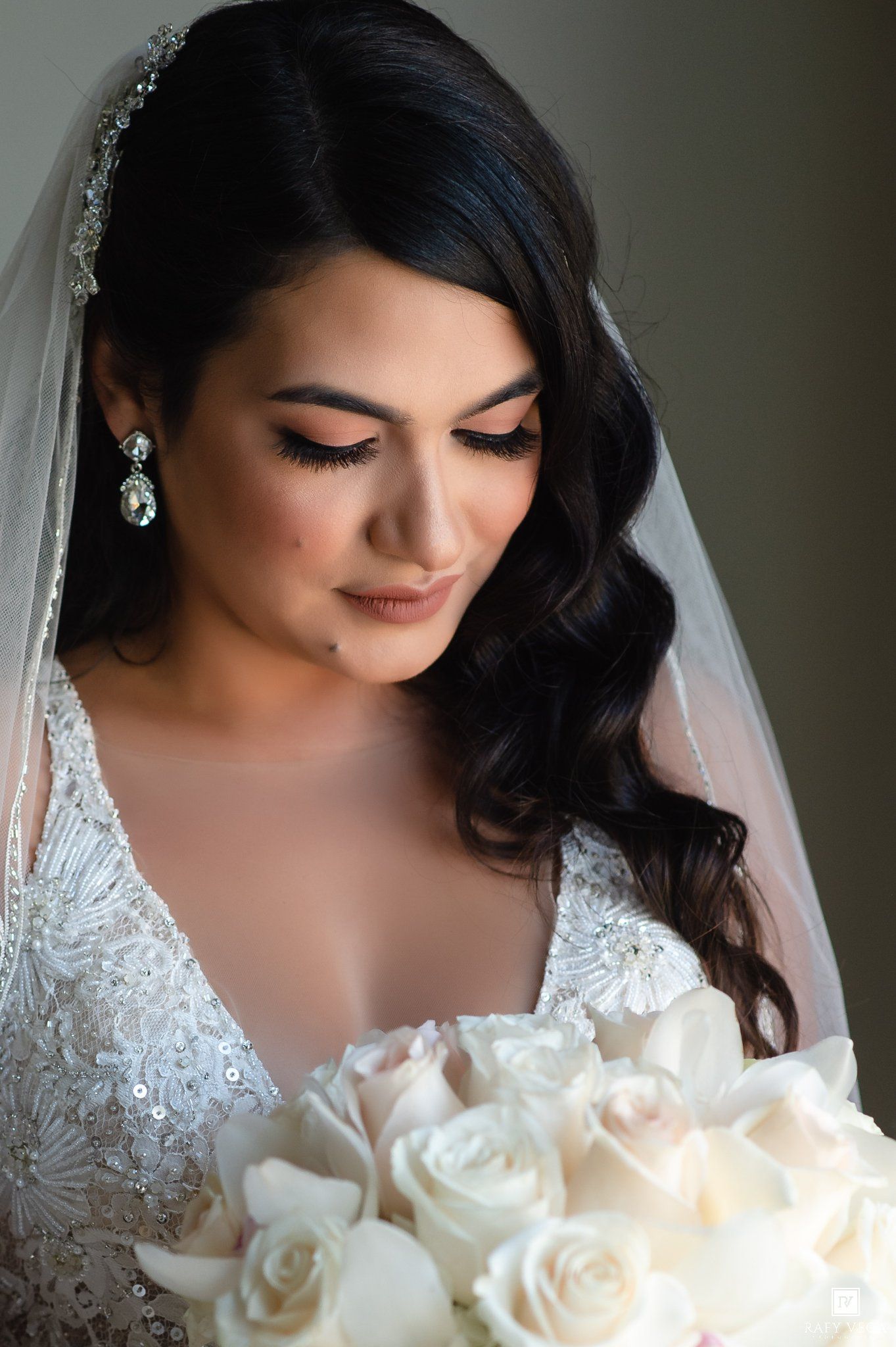 Bride in beaded white gown holds ivory rose bouquet, wearing crystal earrings and veil, looking down gracefully.
