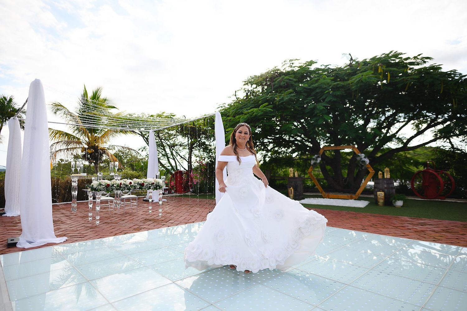 Bride in white wedding gown twirling on outdoor dance floor surrounded by tropical trees and elegant white decor.