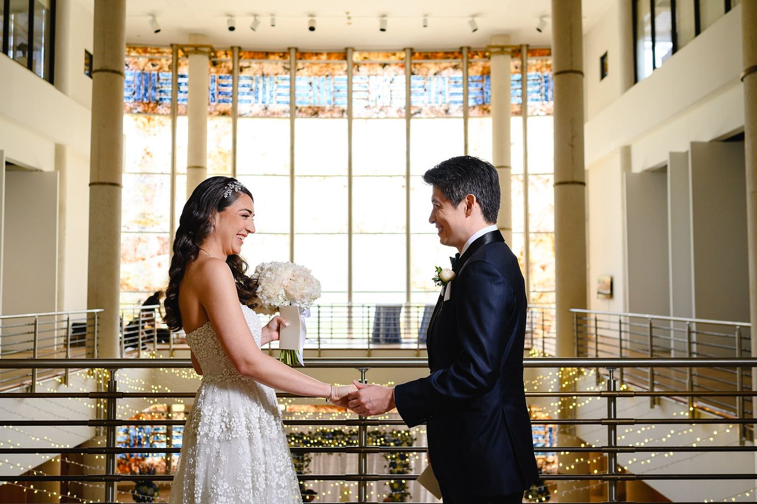 Bride and groom hold hands and smile at each other in an elegant venue with stained glass windows.