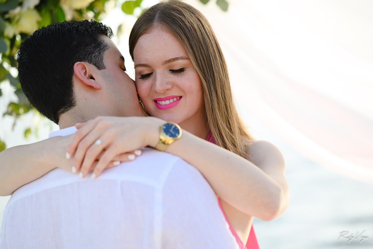 Happy couple embracing outdoors, woman smiling warmly wearing a blue and gold watch, romantic beach setting.
