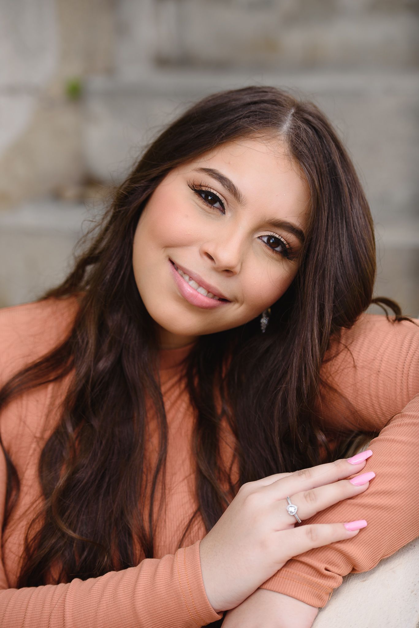 Young woman with long dark hair smiling, wearing a peach top and pink nails, posing casually indoors.