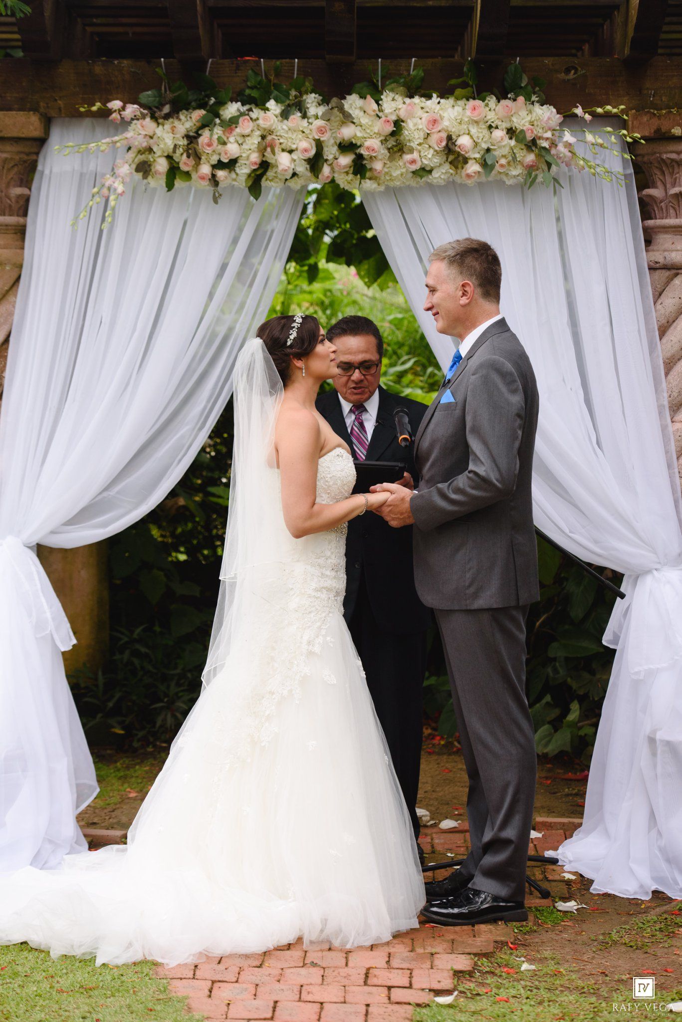 Bride and groom exchanging vows under floral arch with white draping at outdoor garden wedding ceremony.