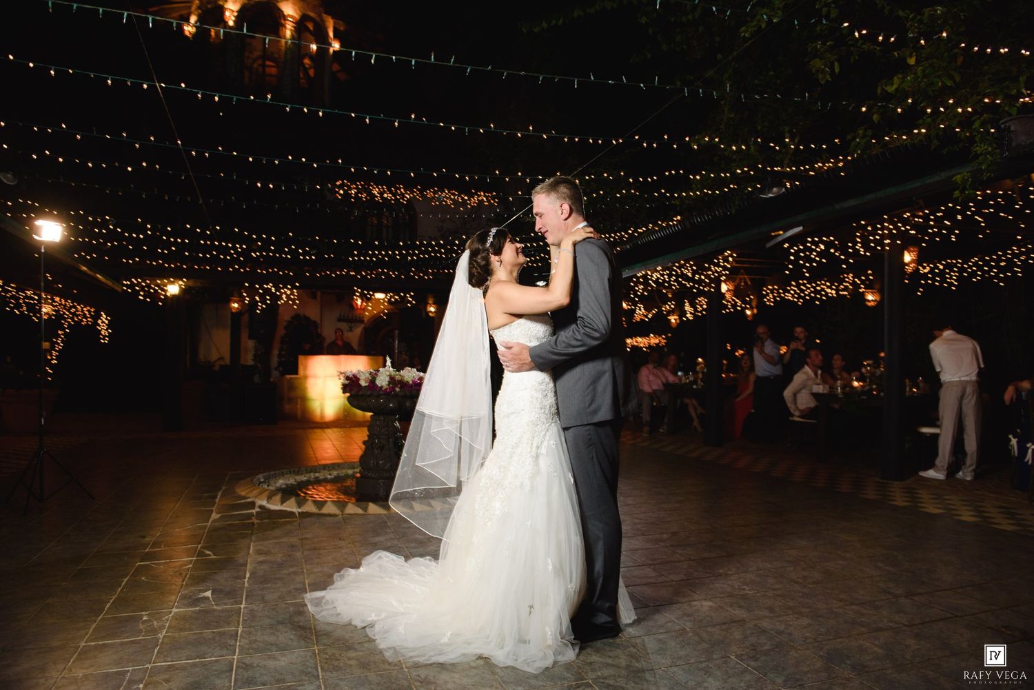 Bride and groom share first dance under twinkling string lights at elegant outdoor wedding reception at night.