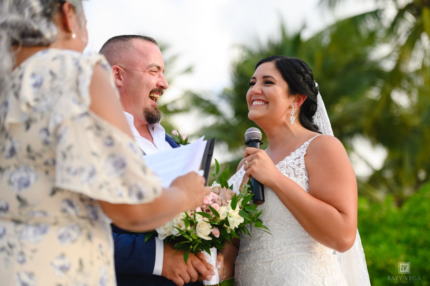 Bride smiling joyfully while reciting wedding vows outdoors, holding bouquet, with groom and officiant at tropical ceremony.