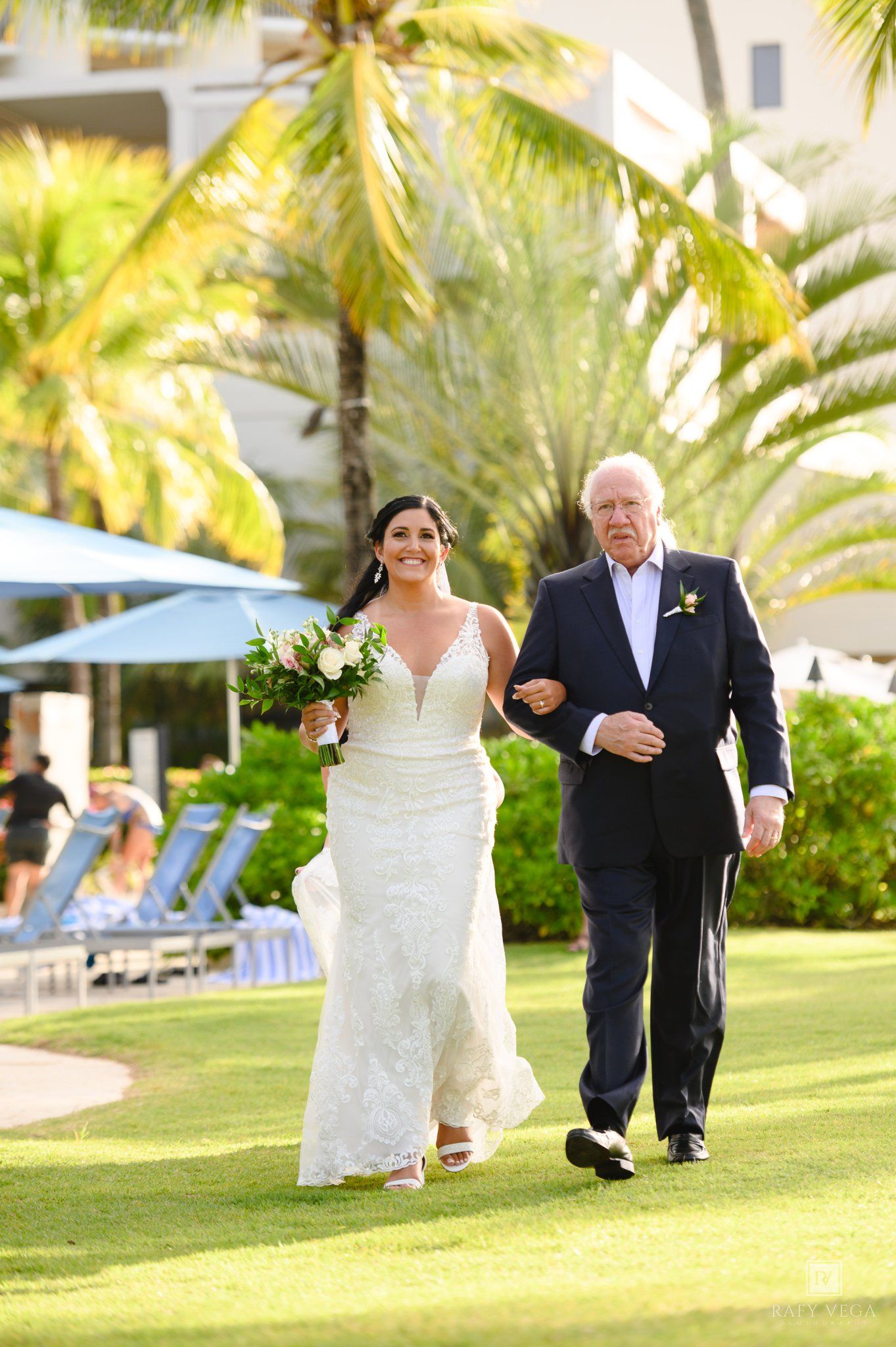 Bride in white lace gown walks with older gentleman in navy suit on green lawn at tropical resort wedding.