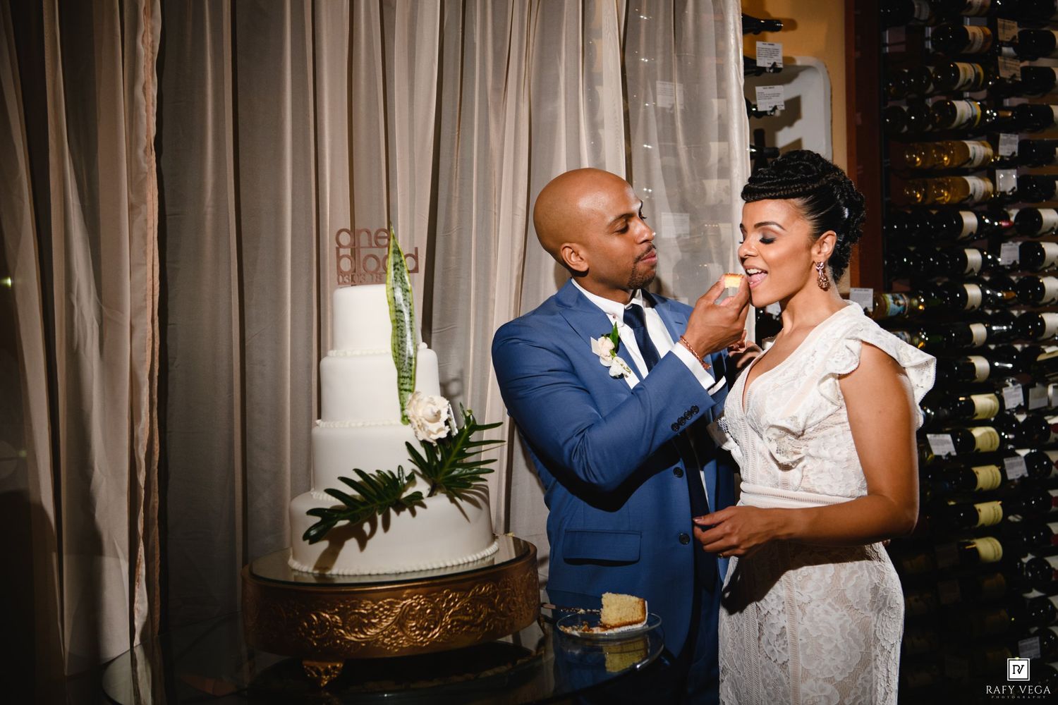 Groom in blue suit feeds wedding cake to bride in elegant white dress near tiered cake display.
