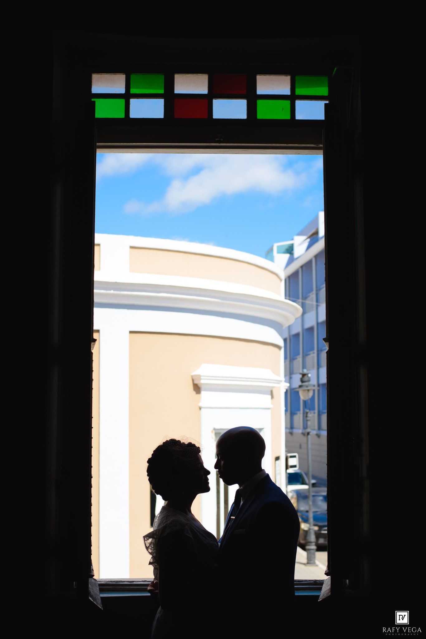 Silhouette of a couple facing each other framed by a doorway with stained glass, blue sky and white building behind.