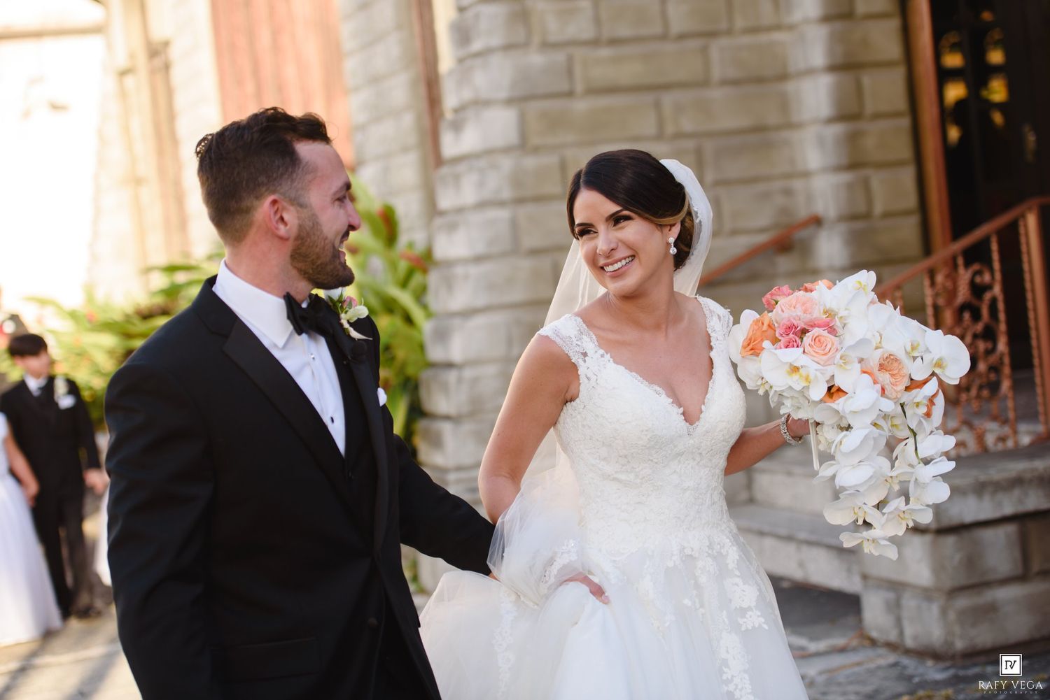 Newlywed couple smiling and holding hands outside a stone church, bride in white gown holding peach and white bouquet.