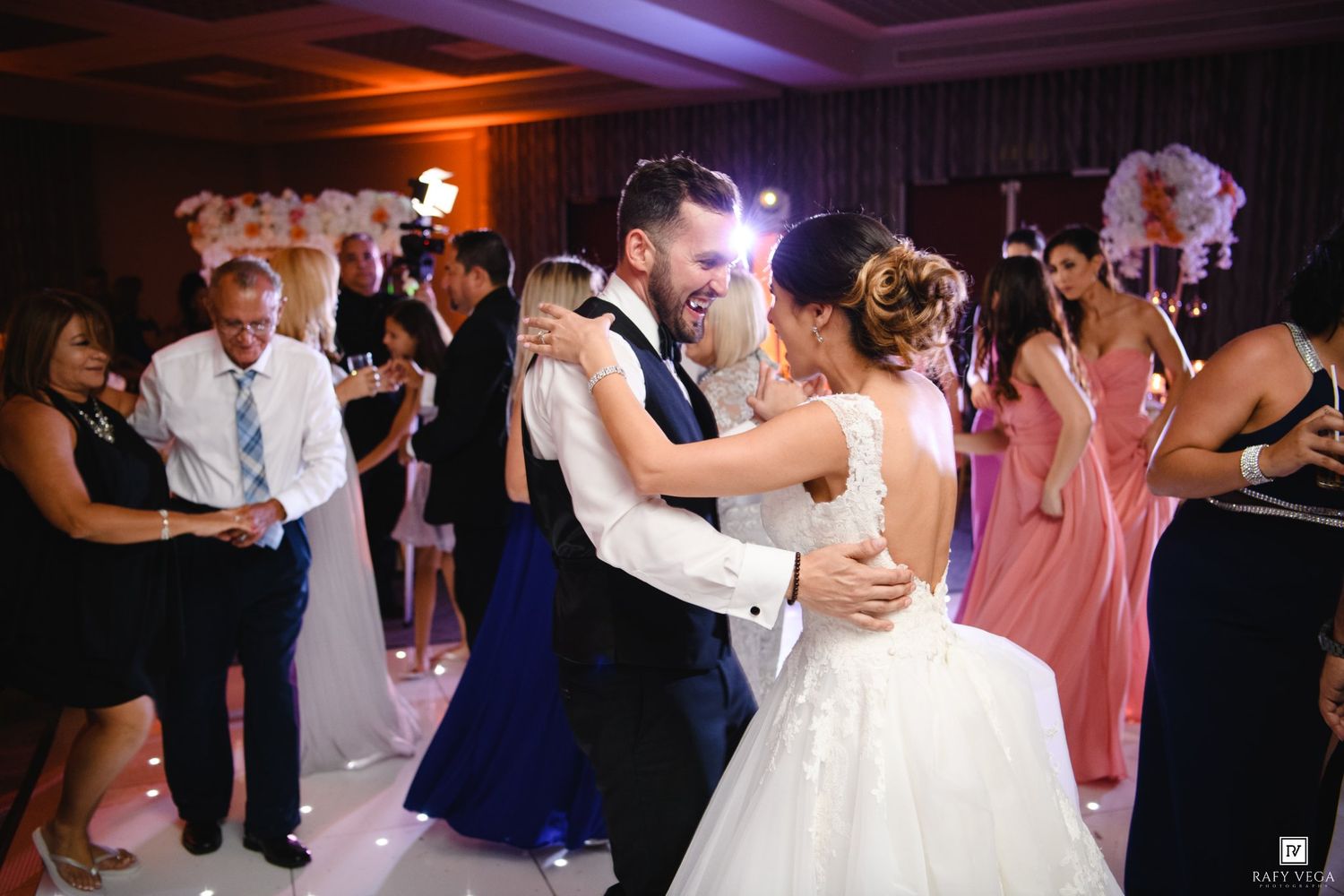 Bride and groom share a romantic first dance embrace at elegant wedding reception with guests and bridesmaids in pink dresses.