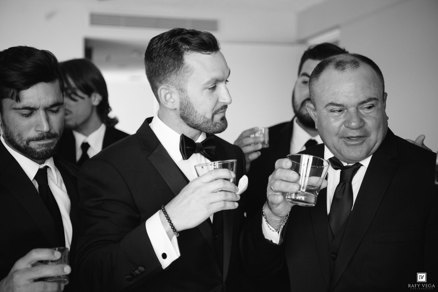 Groom in black tuxedo toasting with groomsmen in black suits during wedding celebration in black and white photo.