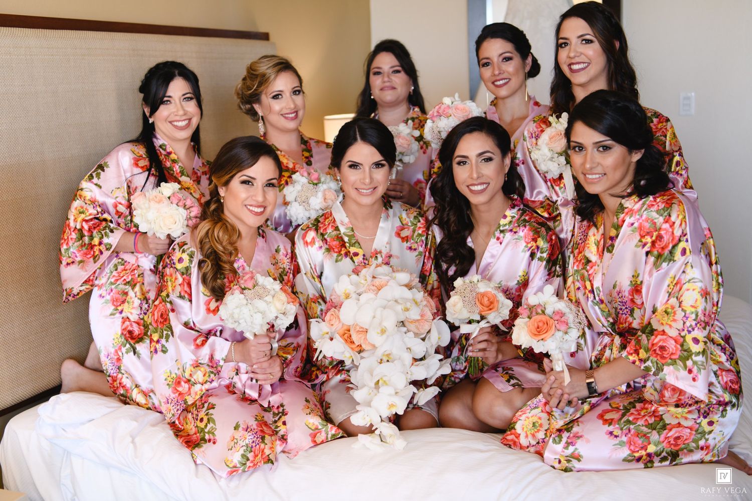 Bridesmaids in matching pink floral robes holding bouquets pose with bride on bed before wedding ceremony.