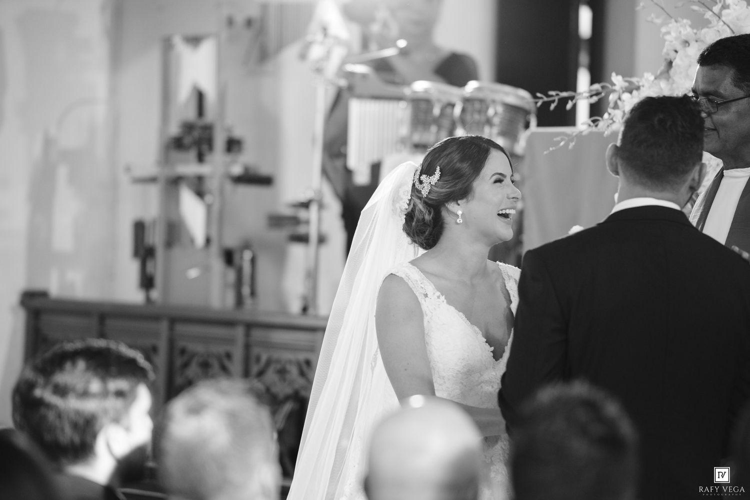 Black and white photo of a smiling bride in veil at church altar during wedding ceremony.