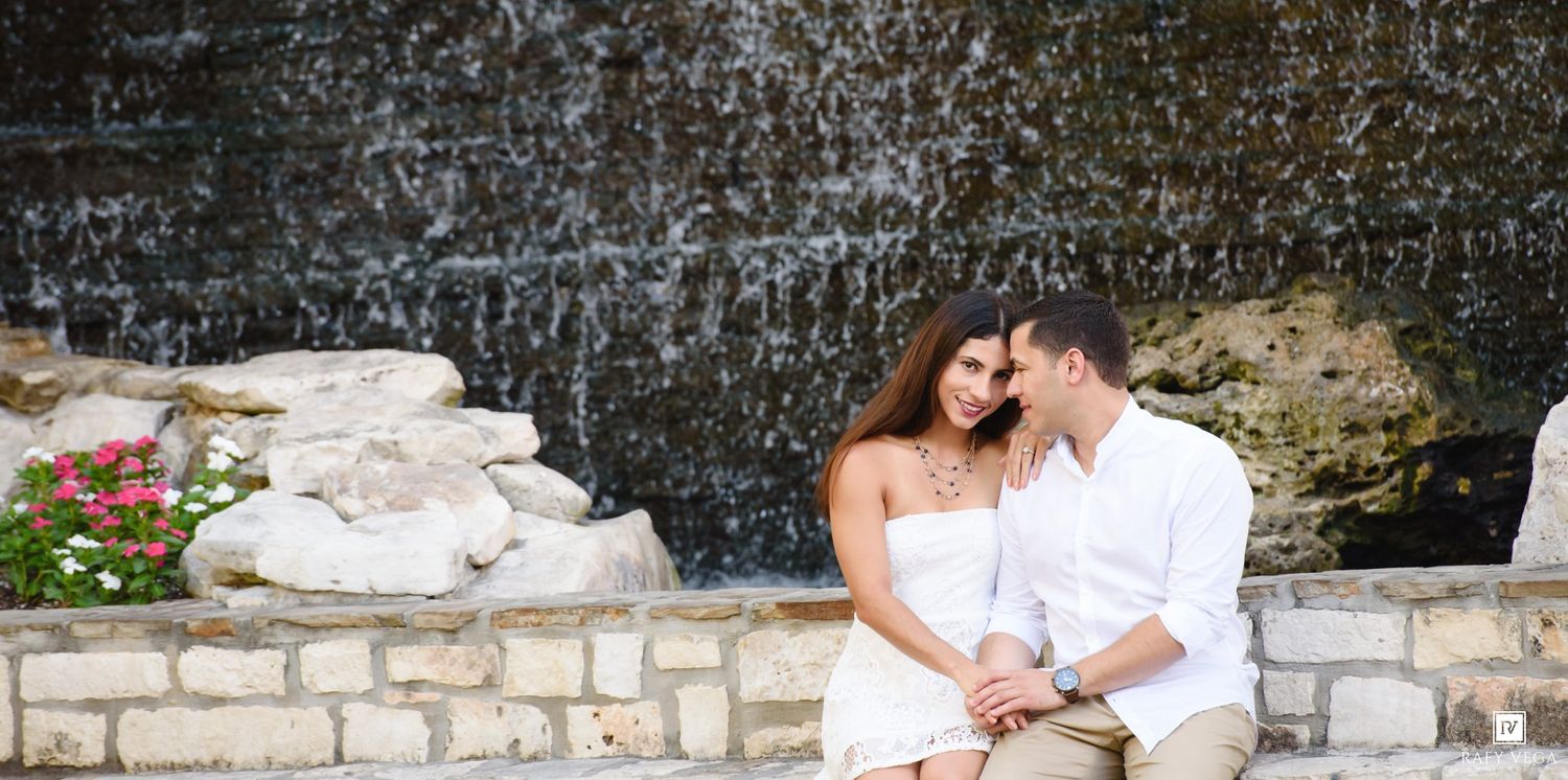 Romantic couple sitting together on stone steps in front of a cascading waterfall surrounded by lush greenery.