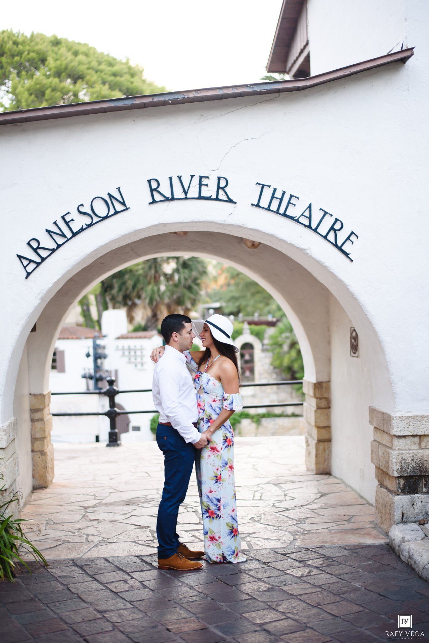 Couple embracing under the Arneson River Theatre arch in San Antonio, TX, in a romantic engagement photo.
