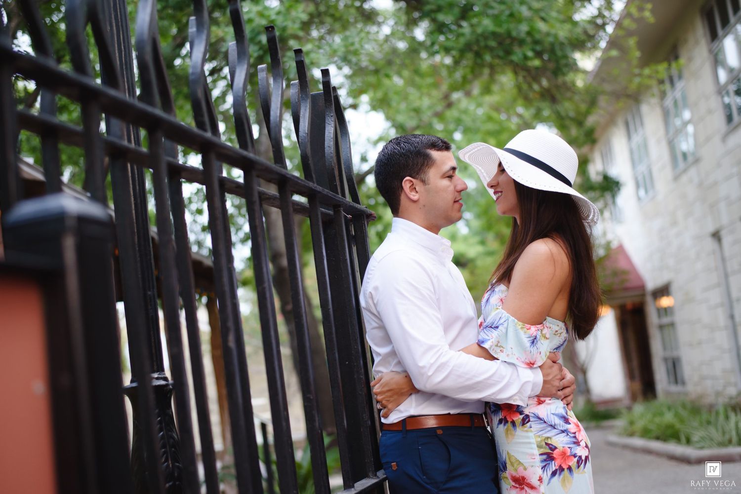 Couple embracing romantically by ornate black iron gate, woman in floral dress and white hat, man in white shirt.