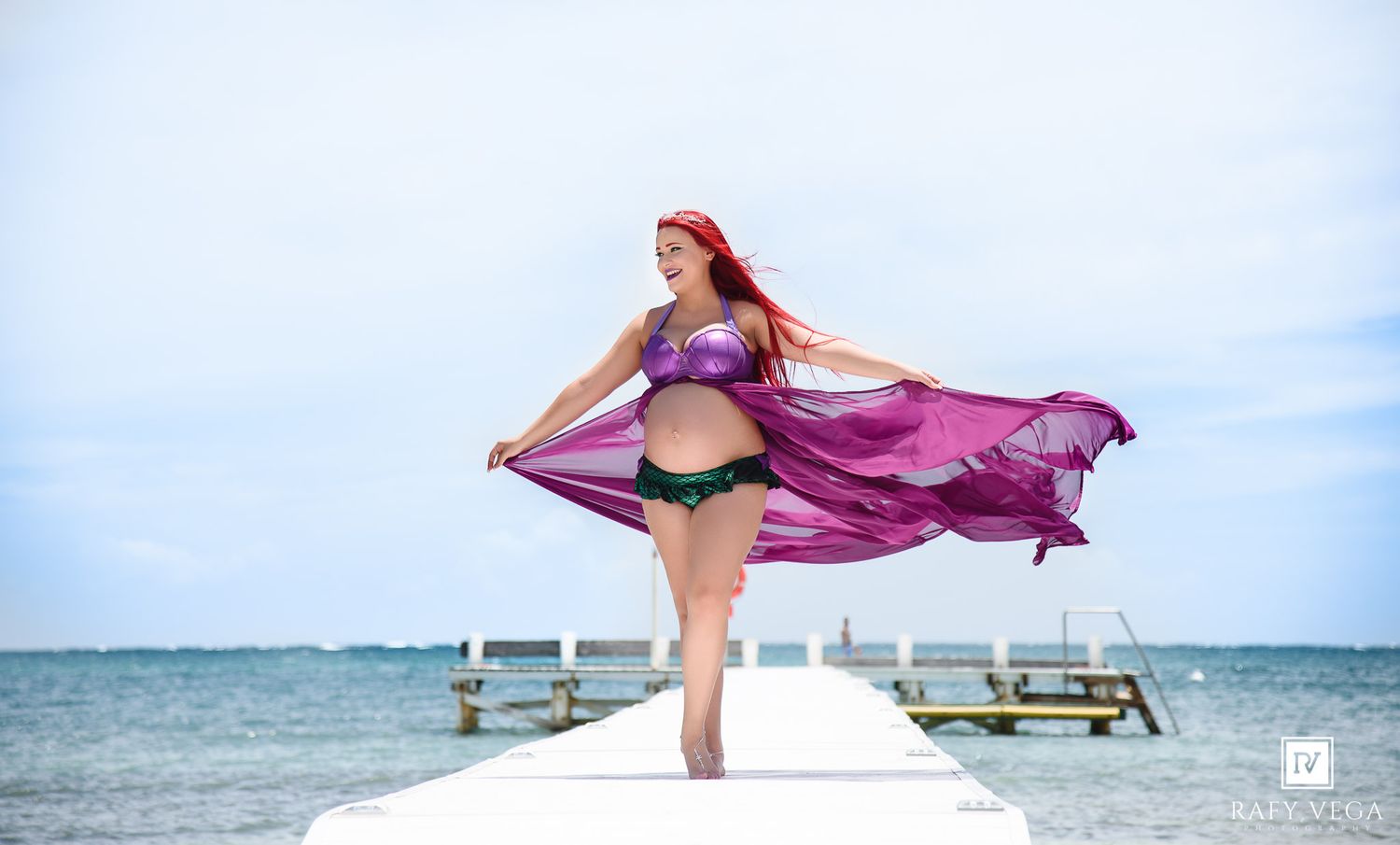 Pregnant woman in mermaid costume with purple flowing fabric poses on a dock by the ocean on a sunny day.