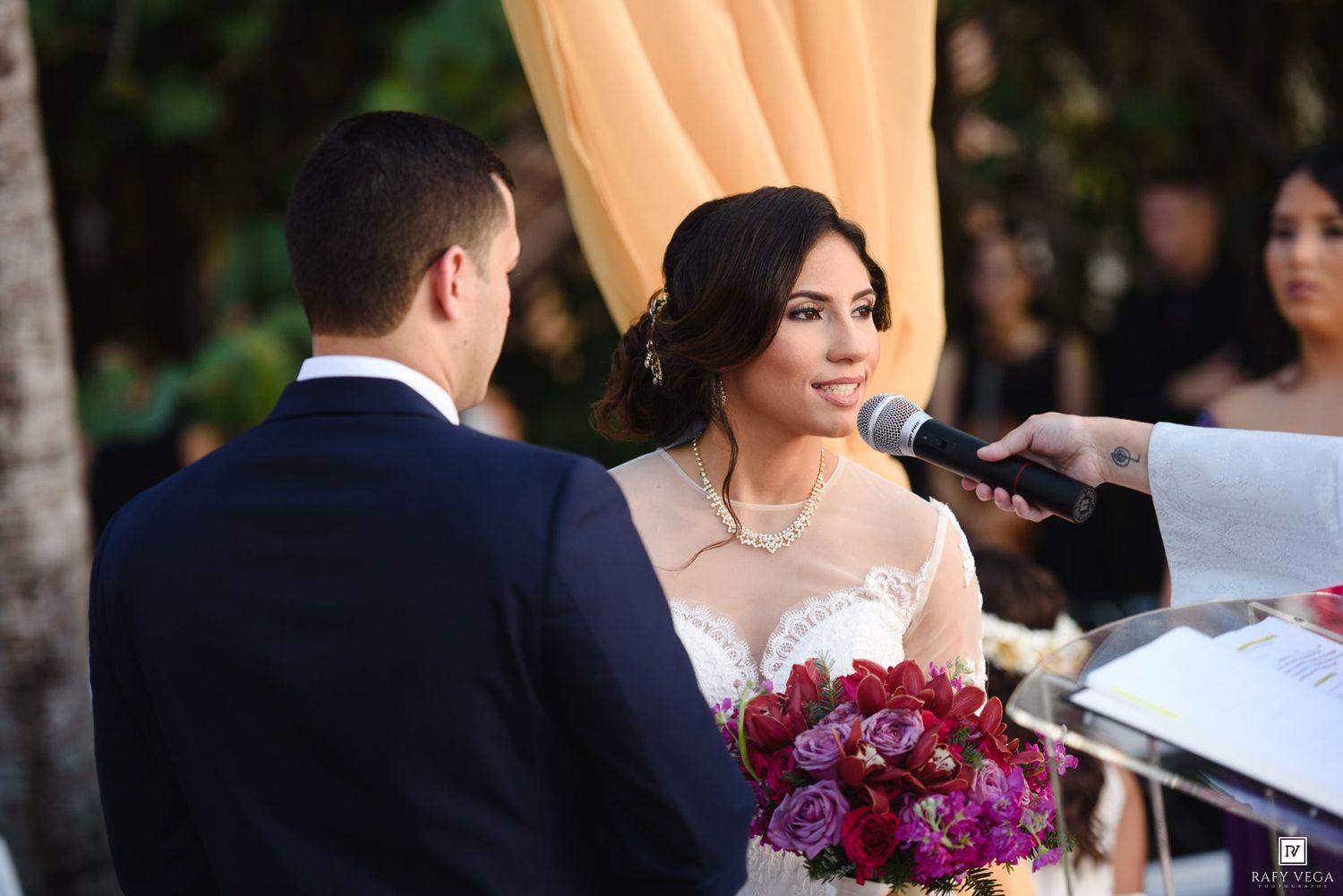 Bride holding vibrant red and purple bouquet recites vows at outdoor wedding ceremony with groom in navy suit.