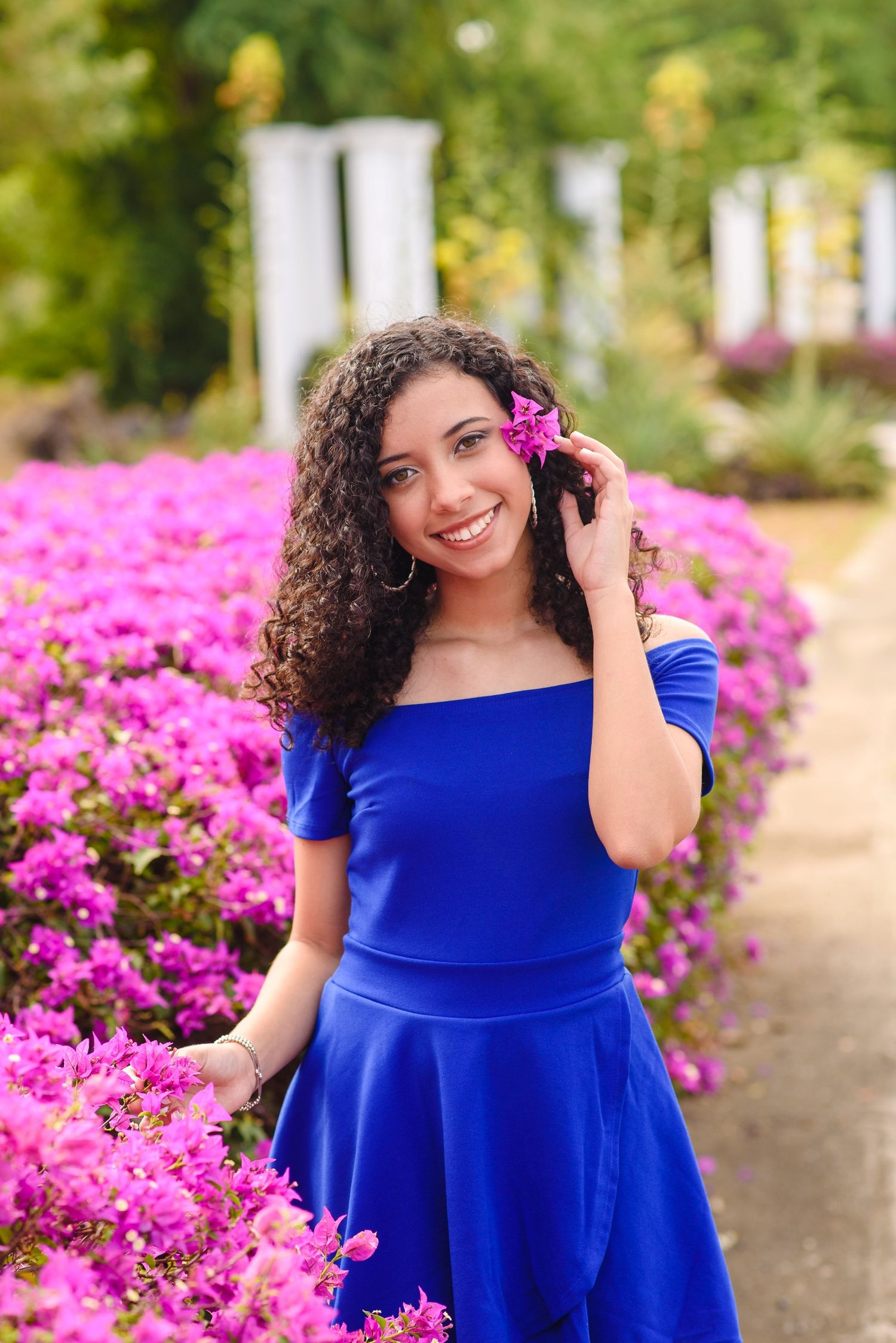 Young woman in blue off-shoulder dress smiling among vibrant pink flowers, holding a bloom near her face.