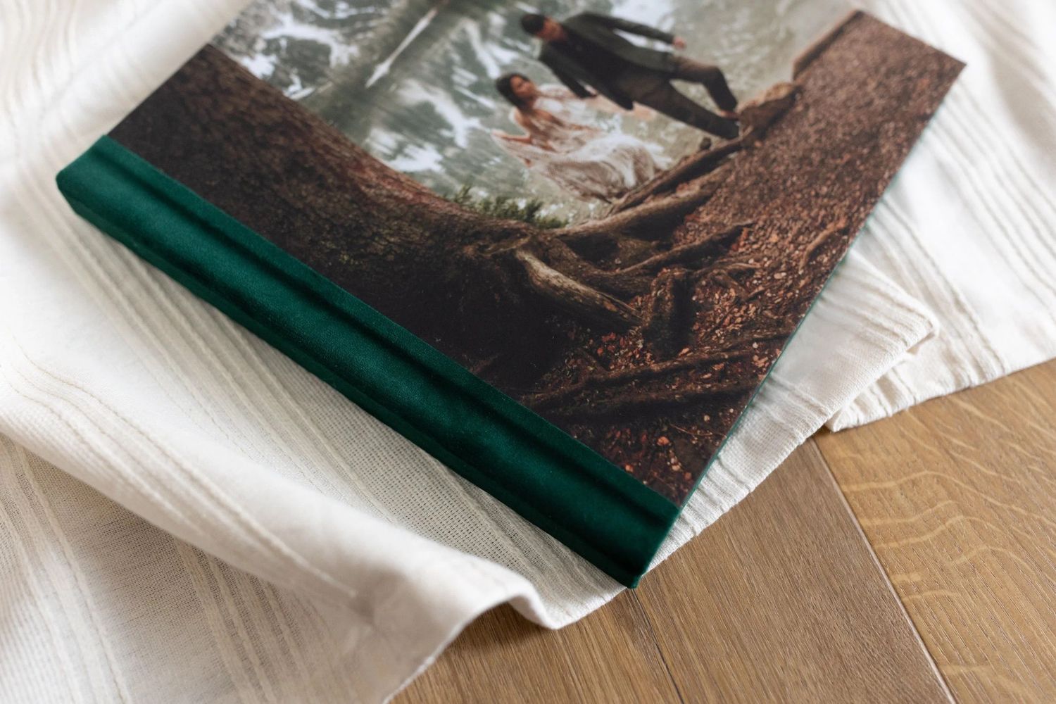 Elegant photo book with green spine and forest wedding cover photo resting on white fabric on wooden surface.