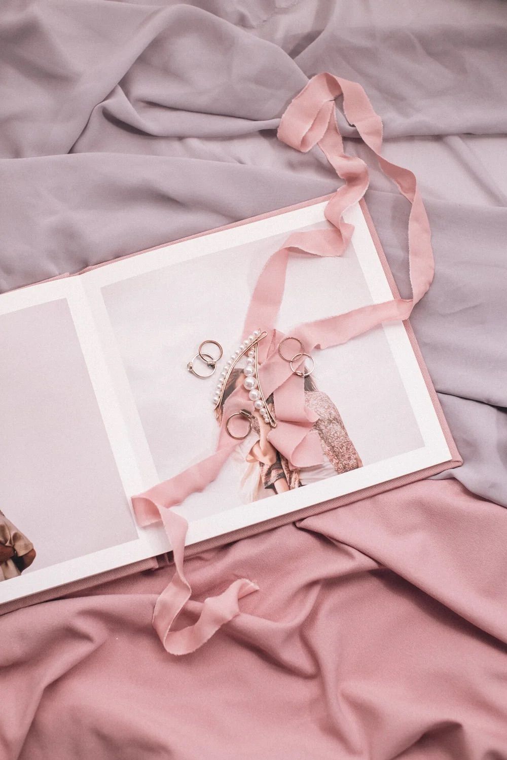 Open wedding photo album with pink ribbon and jewelry on soft pink and gray fabric backdrop.