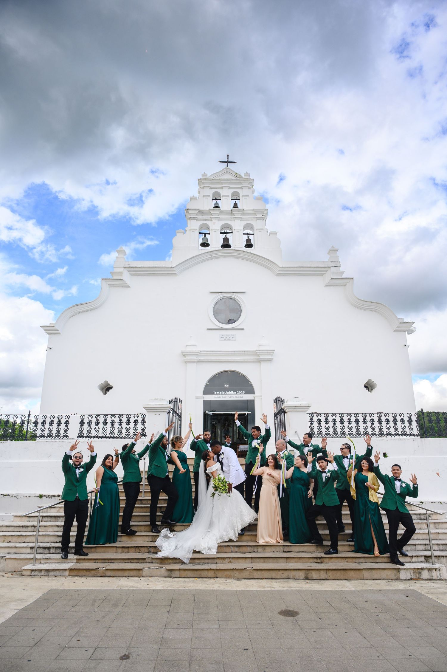 Wedding party celebrating on steps of white church with green bridesmaid dresses and groomsmen suits under cloudy sky.