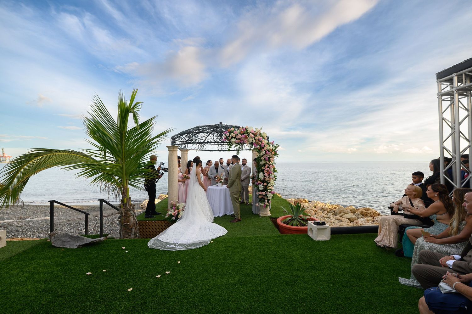 Outdoor beach wedding ceremony with couple at floral arch, palm tree, green grass, and ocean backdrop under a blue sky.