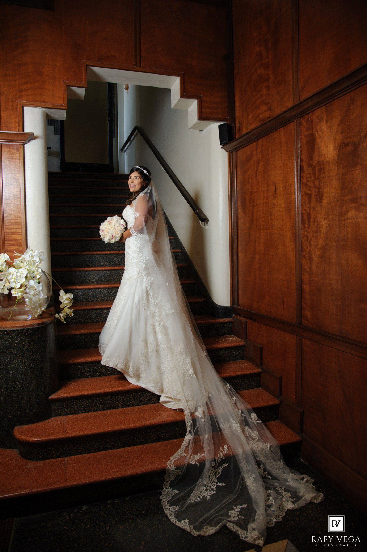 Bride in lace mermaid wedding gown with cathedral veil poses on elegant wooden staircase holding white bouquet.