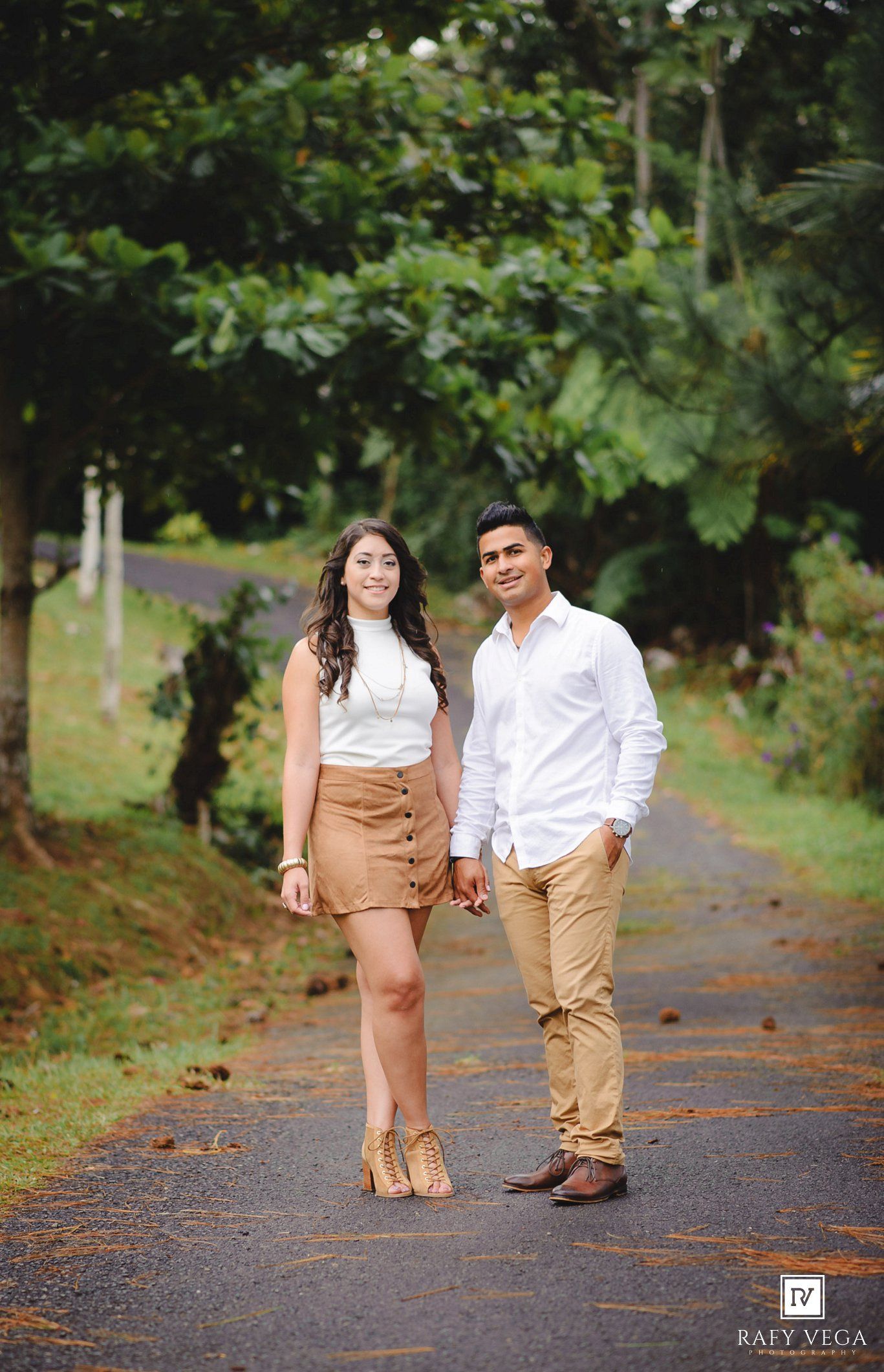 Couple posing on a lush green tree-lined path, woman in white top and tan skirt, man in white shirt and khaki pants.