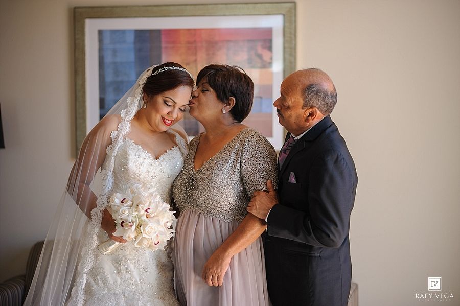 Bride in lace wedding gown receives a kiss from her mother while her father looks on lovingly.