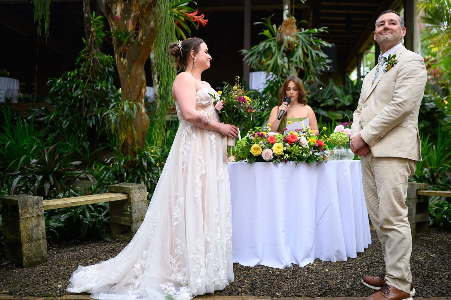 Bride in lace gown and groom in tan suit exchange vows outdoors at a tropical garden wedding ceremony.