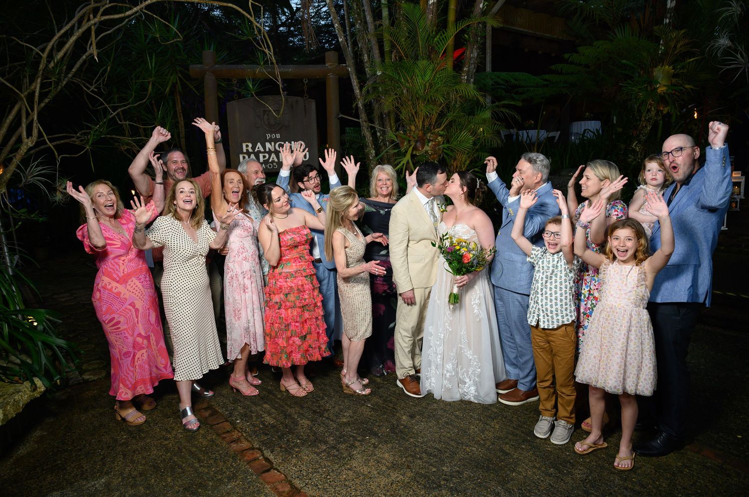 Wedding group photo with bride and groom kissing surrounded by cheering family and friends outdoors at night.