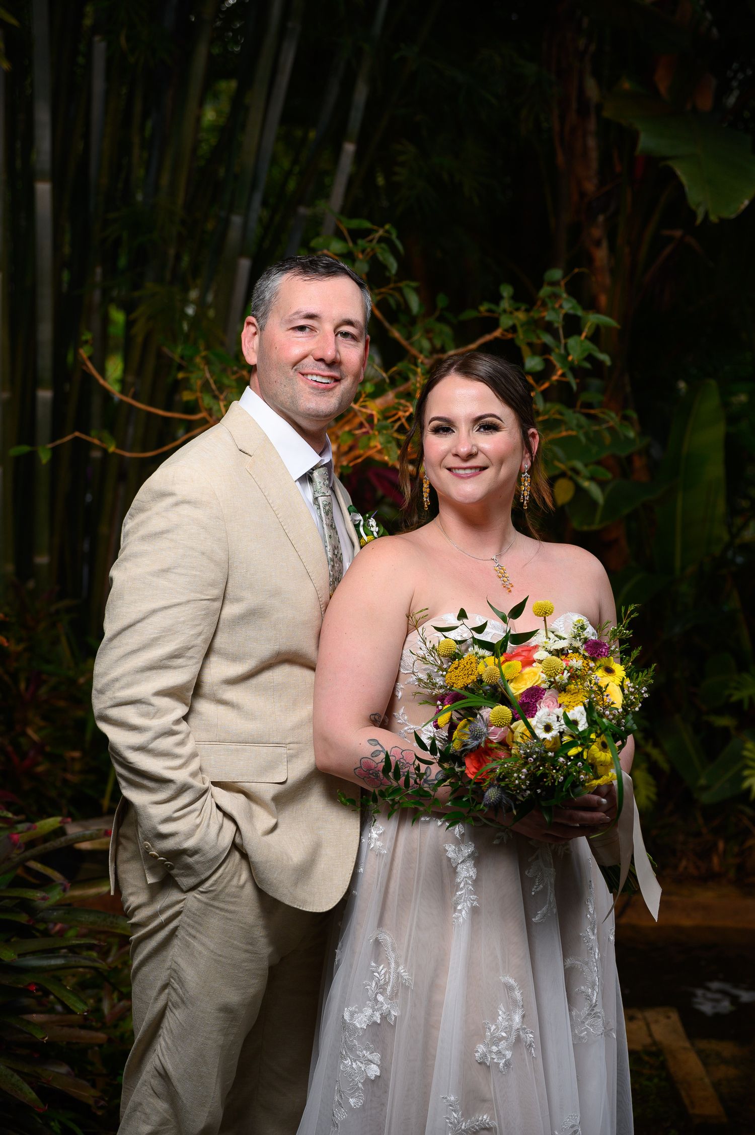 Smiling bride and groom pose together in tropical garden setting, bride holding colorful bouquet in white lace gown.