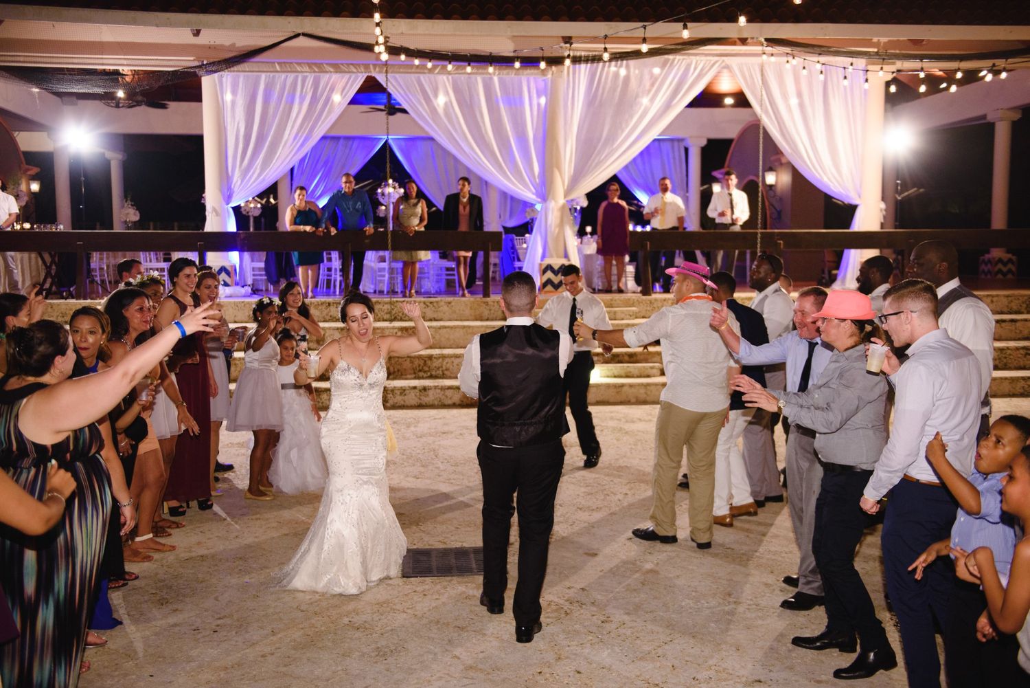Bride and groom enter wedding reception as guests cheer under elegant white draped canopy with purple lighting.