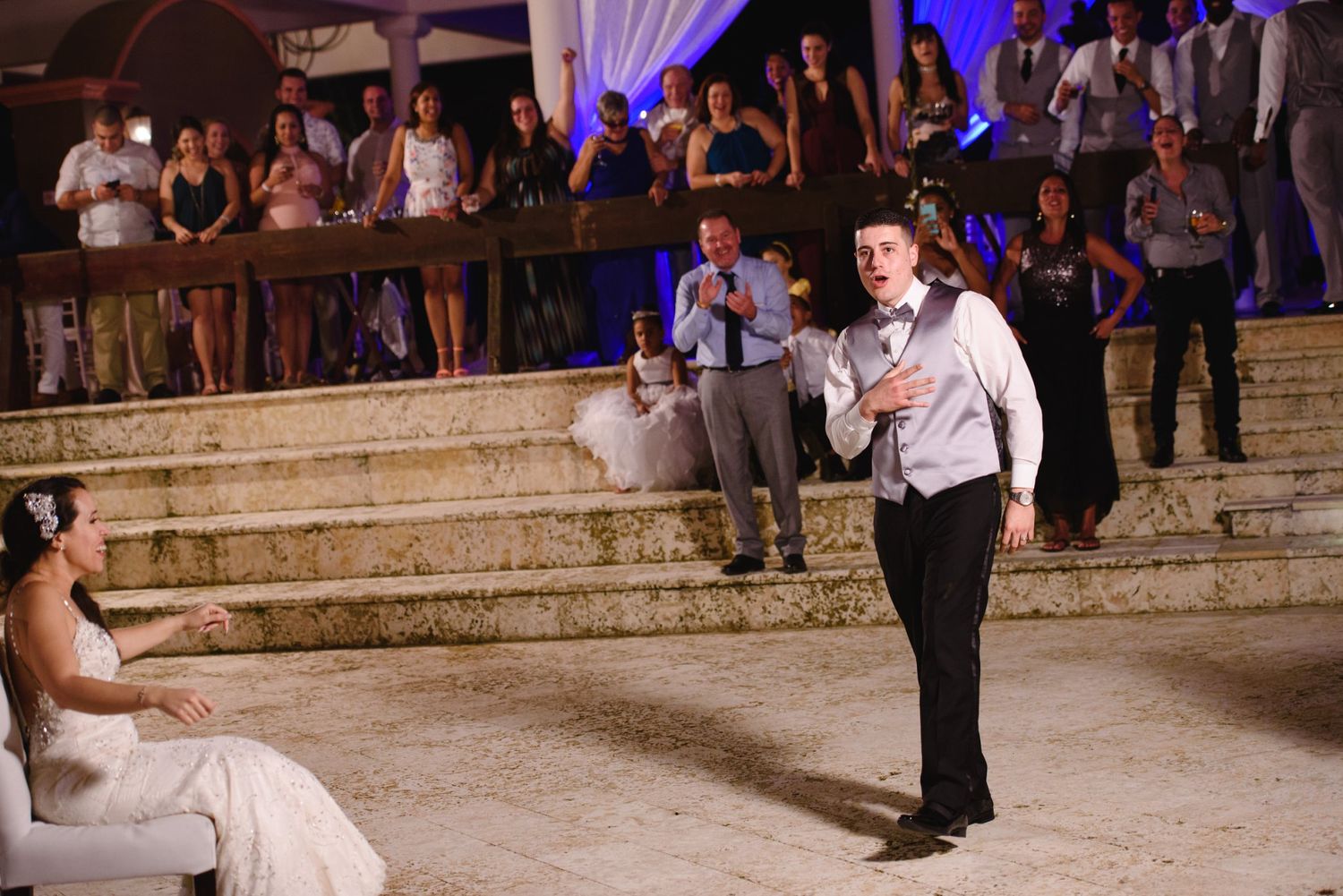 Groom serenading bride at outdoor wedding reception while guests watch from stone steps.