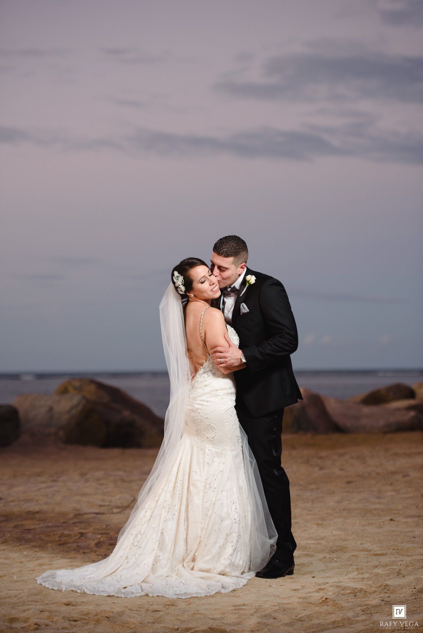 Bride and groom share a romantic embrace on a sandy beach at sunset, with the bride in a lace gown and veil.
