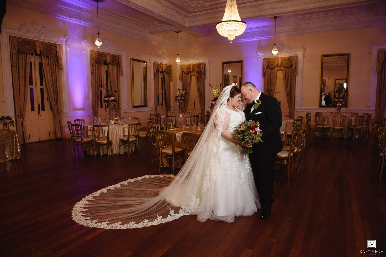 Bride and groom share a romantic kiss on the dance floor of an elegant ballroom decorated with purple uplighting.