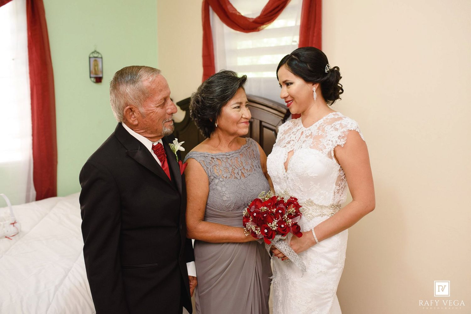 Bride in lace gown holds red bouquet with parents in elegant room before wedding ceremony.
