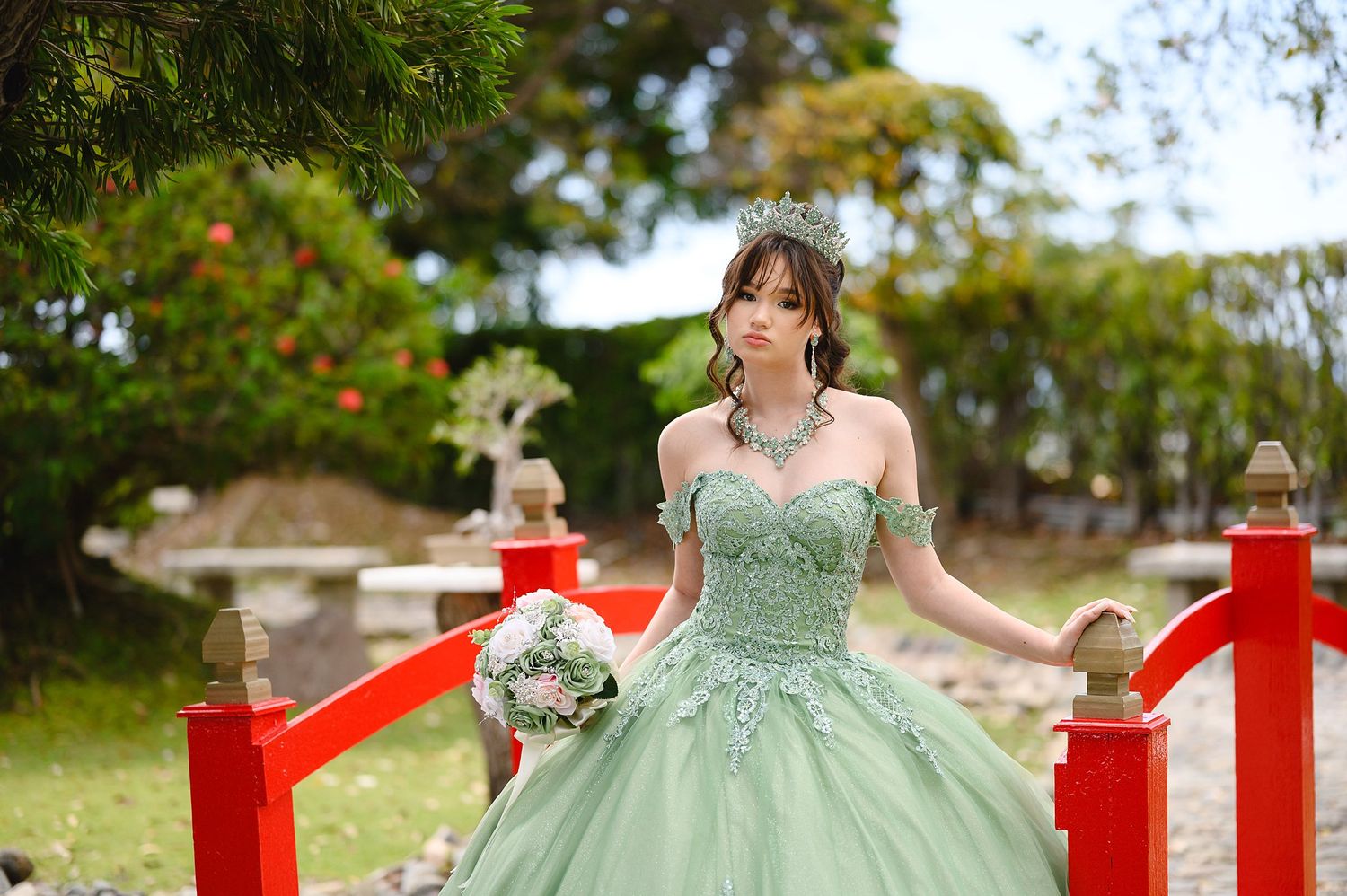 Young woman in mint green off-shoulder ball gown with crown and bouquet poses on red bridge in garden.
