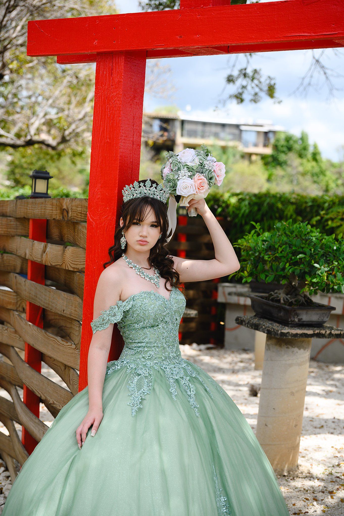 Young woman in sage green quinceañera ball gown with crown and floral bouquet poses by red torii gate.