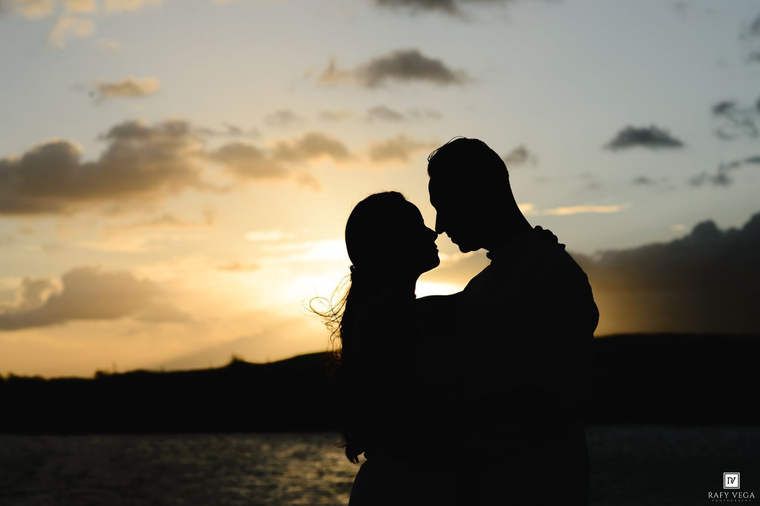 Silhouette of a romantic couple about to kiss at sunset near water with dramatic cloudy sky backdrop.