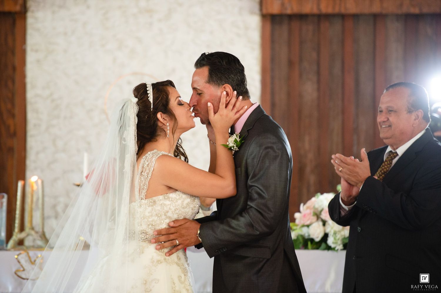 Bride and groom share first kiss at wedding ceremony altar while officiant applauds in elegant venue.