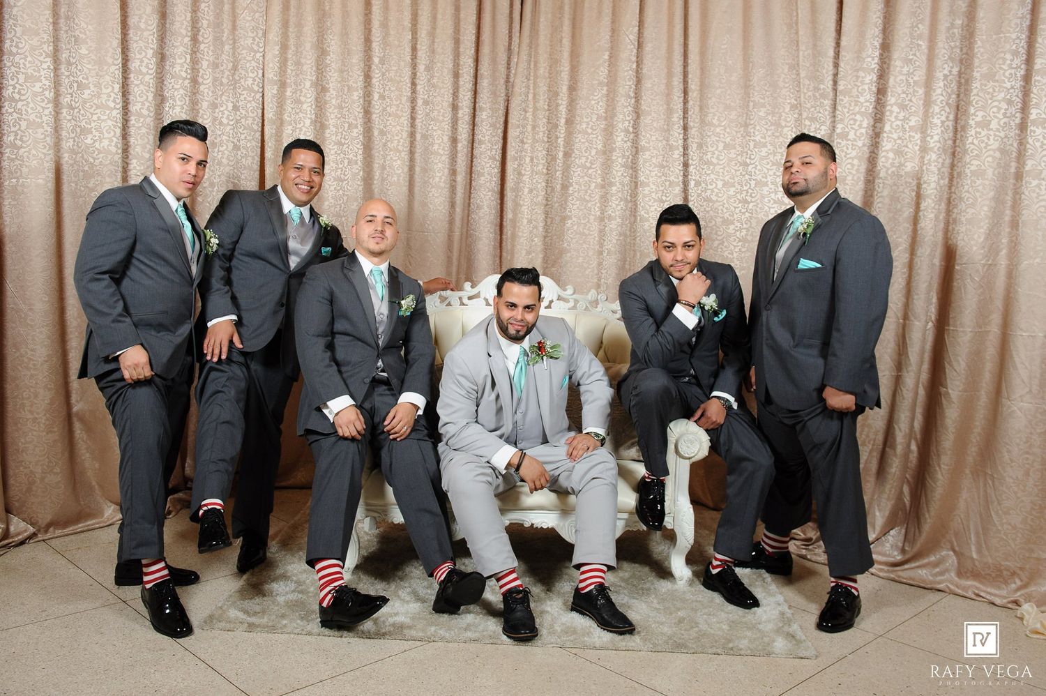 Groom in light gray suit sits on white throne chair surrounded by groomsmen in dark gray suits with green ties.