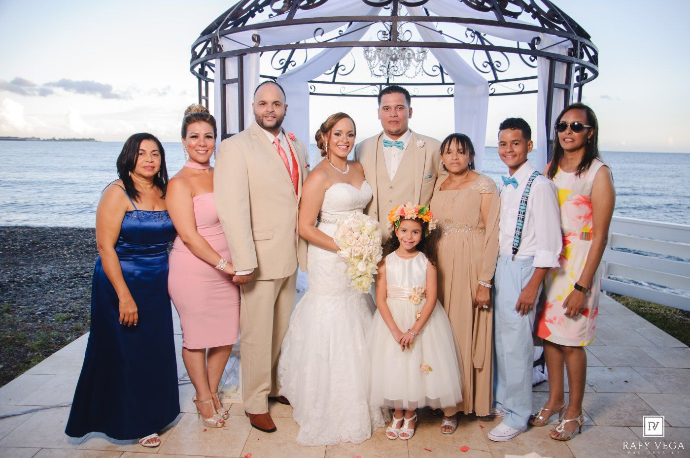 Wedding party group portrait with bride, groom, family and flower girl posed in front of beachside gazebo at sunset.