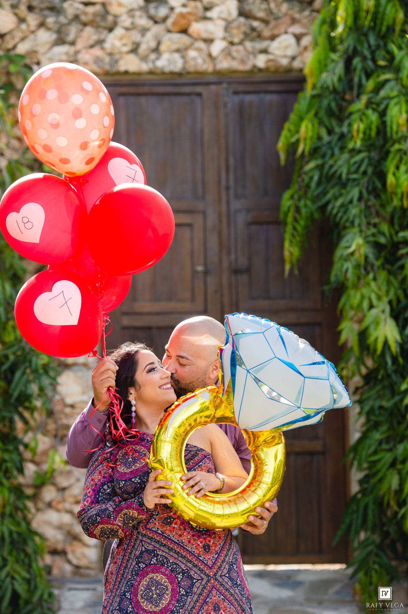 Couple celebrating engagement with red heart balloons and diamond ring balloon in front of rustic stone wall.